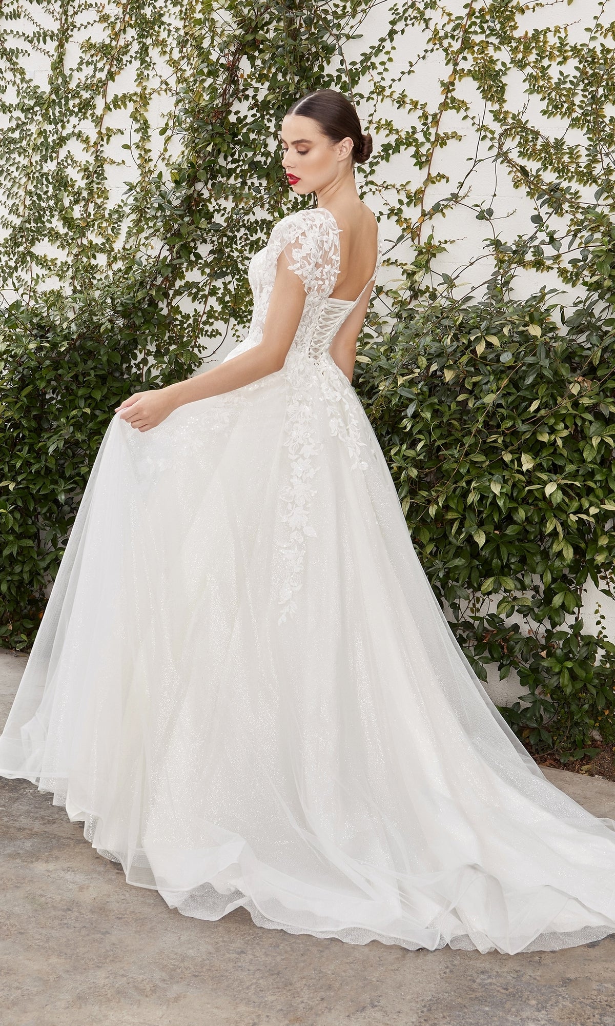 A woman wears the Cap-Sleeve White Wedding Gown: Ladivine A1082W, featuring floral lace, a fitted bodice, and flowing train, as she stands outdoors near a vine-covered wall.